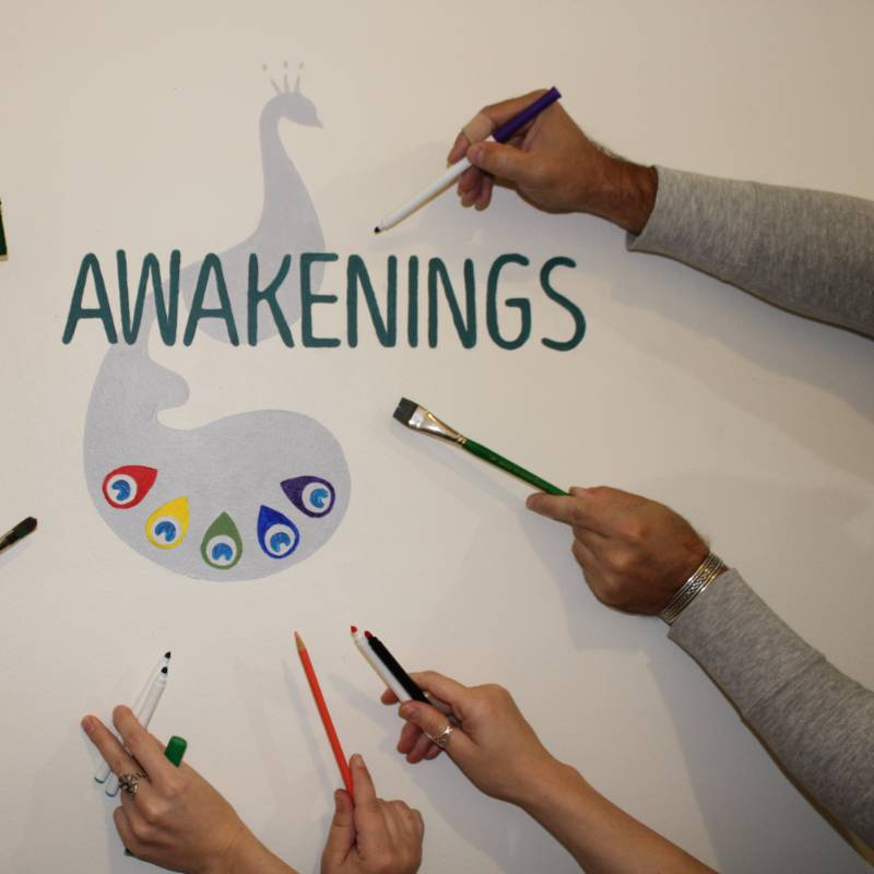 Three hands holding drawing and painting tools point toward a wall mural featuring the word "AWAKENINGS" and a stylized peacock design.
