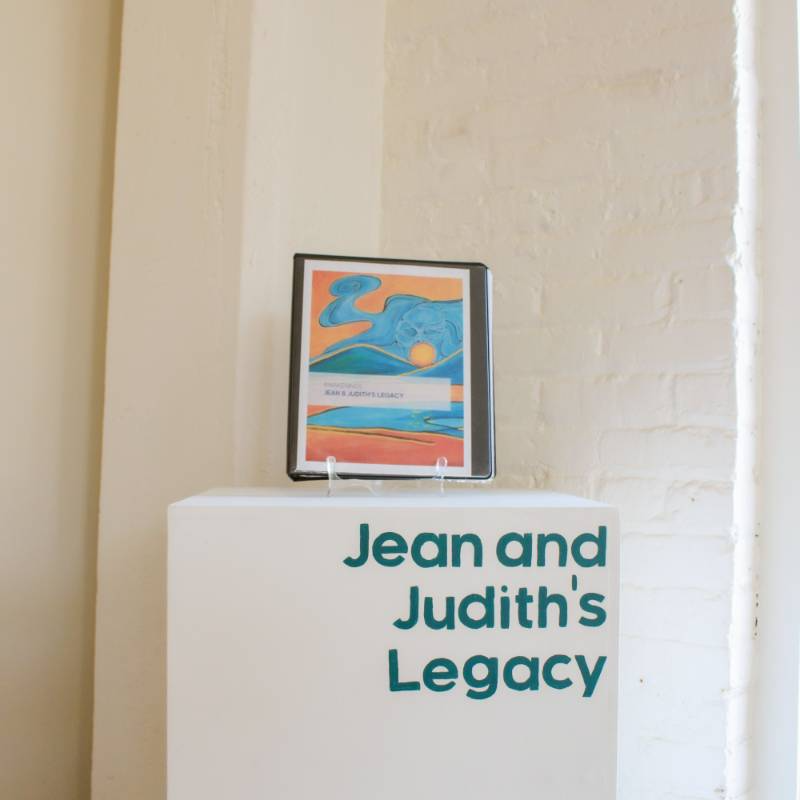A display pedestal labeled "Jean and Judith's Legacy" holds a binder with an abstract cover featuring blue and orange artwork.