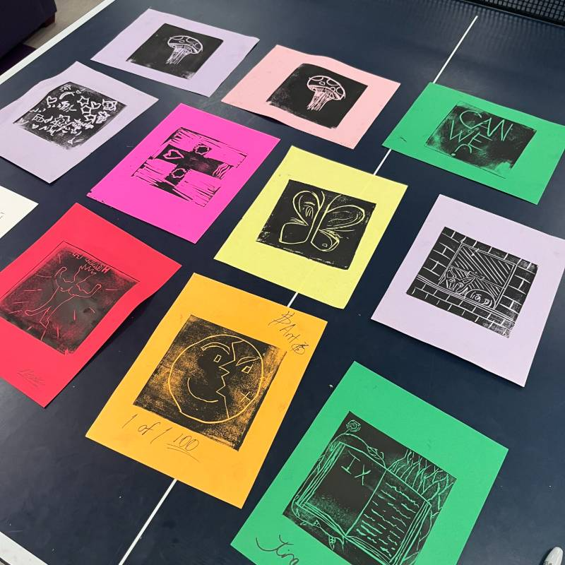 Twelve colorful paper prints with various abstract and figurative black ink designs are arranged on a table in a grid pattern.