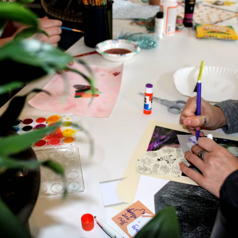 People working on art and craft projects at a table with supplies including watercolor paints, glue sticks, scissors, colored pencils, and paper.