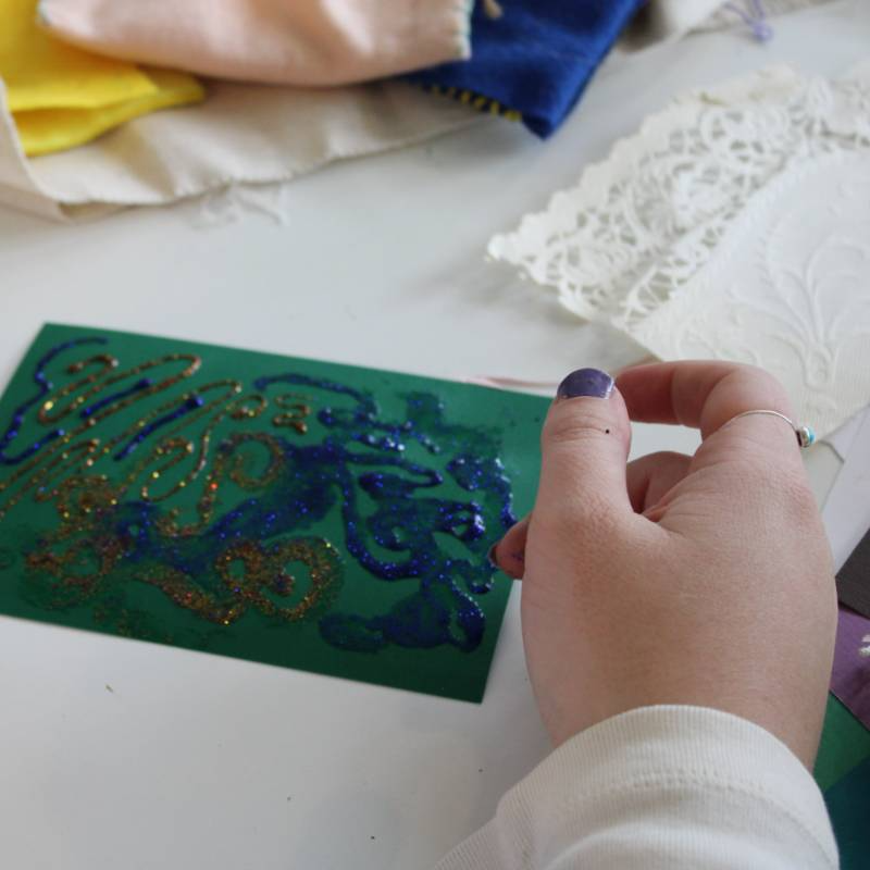A person with a purple-stained thumb holds a green card decorated with blue and gold glitter, surrounded by various fabric and paper materials on a table.