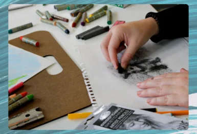 A person uses charcoal on sketch paper, with art supplies such as oil pastels, drawing board, and packaging scattered on a white table.