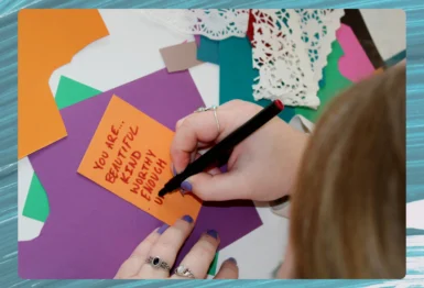 A person writes “You are: beautiful, valid, worthy, enough, unique” on an orange card surrounded by colorful paper and craft supplies.