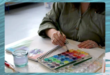 A person uses a brush to paint in a sketchbook with a watercolor palette, glass of water, and art supplies on the table.