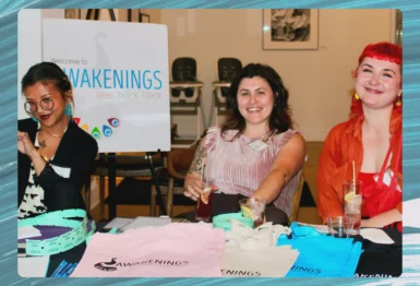 Three people sit at a table with drinks and Awakenings tote bags in front of a sign that reads "Awakenings See Hear Heal" in a well-lit indoor space.