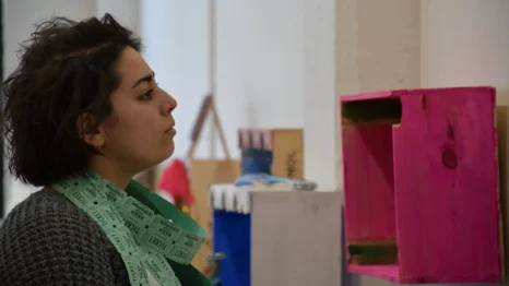 A person with dark hair and a bundle of green tickets around their neck looks at a pink display shelf in an indoor setting.