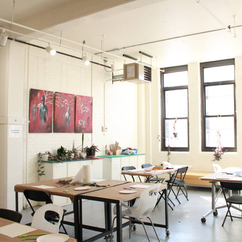 Bright art classroom with tables, chairs, art supplies, and two red paintings on the wall; sunlight streams through large windows.