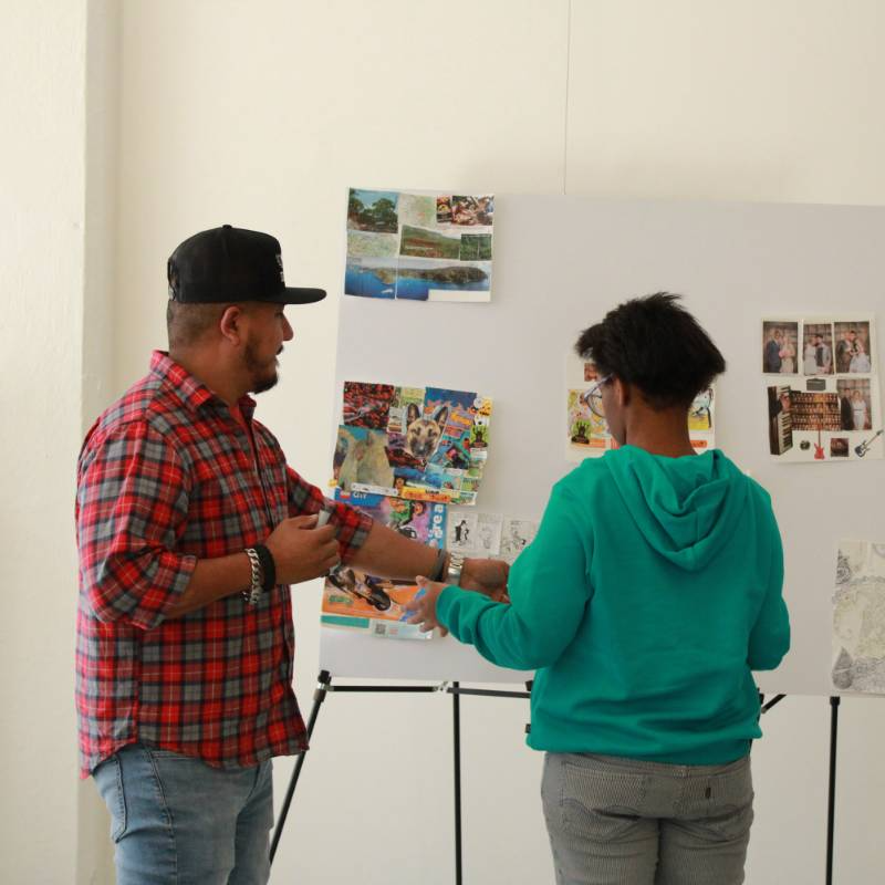 Two people stand in front of a display board covered with photos and images, discussing or arranging the items on the board.