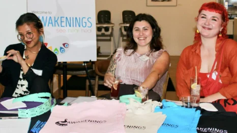 Three women sit at a table with colorful Awakenings tote bags and paperwork, smiling. A welcome sign with “Awakenings” is visible in the background.