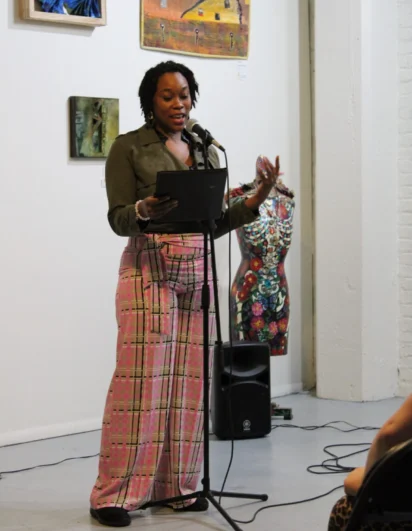 A woman stands at a microphone reading from a tablet in an art gallery, with colorful artworks displayed on the wall behind her.