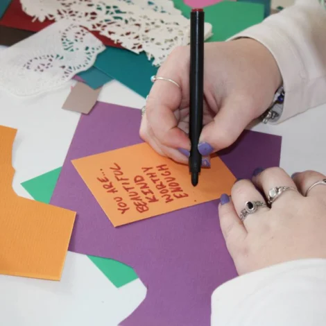 A person writes a positive message on an orange card surrounded by colorful craft supplies and paper shapes.