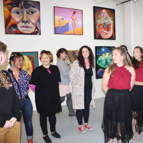 A group of people stand and talk in an art gallery with colorful paintings displayed on white walls behind them.