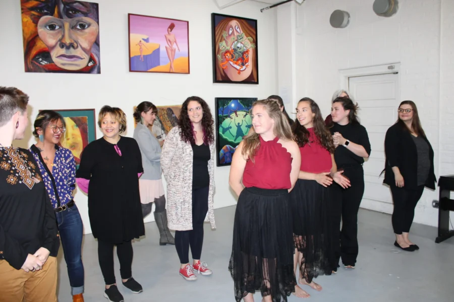 A group of people stand and talk in an art gallery, with colorful paintings displayed on the white walls behind them.