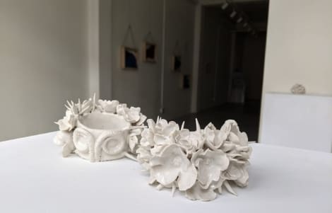 Two white ceramic sculptures featuring floral and spike designs are displayed on a white pedestal in an art gallery setting.