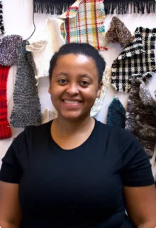 Bryana Bibbs smiles while standing in front of a fiber arts piece with various colors and types of fabrics tied into bows. Bryana is a Black woman with a big smile and hair pulled back.