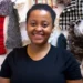 Bryana Bibbs smiles while standing in front of a fiber arts piece with various colors and types of fabrics tied into bows. Bryana is a Black woman with a big smile and hair pulled back.