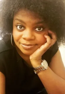 Christina Bourne is a Black woman with an afro, wearing a black top, and leaning her head on her hand while looking at the camera.