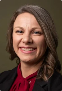 Darci Flynn has curled brown hair, is wearing a red blouse under a black blazer, and is smiling while standing in front of a brown backdrop.