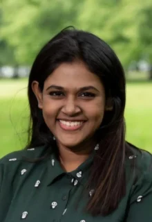 Deya Chatterjee has dark brown long hair, is wearing a dark green collared blouse, and is smiling at the camera while outside in a green space.
