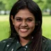 Deya Chatterjee has dark brown long hair, is wearing a dark green collared blouse, and is smiling at the camera while outside in a green space.