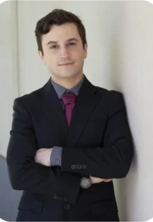 Geetho El Masri is standing with his arms crossed, leaning against a wall, and is wearing a sharp black suite, with a gray collared shirt, and maroon tie.