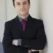 Geetho El Masri is standing with his arms crossed, leaning against a wall, and is wearing a sharp black suite, with a gray collared shirt, and maroon tie.