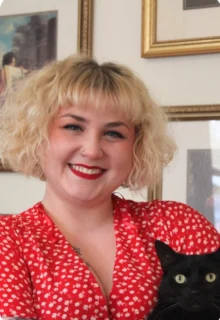 Gillian Marwood has a curly blonde bob, is wearing a red polka-dot dress, and holding a black cat while smiling at the camera.