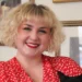 Gillian Marwood has a curly blonde bob, is wearing a red polka-dot dress, and holding a black cat while smiling at the camera.