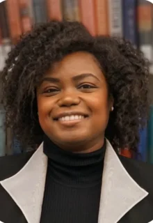 Holley Bloomfield is a Black woman with curly hair wearing a black turtleneck under a collared blazer, smiling at the camera in front of a row of books.