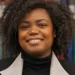 Holley Bloomfield is a Black woman with curly hair wearing a black turtleneck under a collared blazer, smiling at the camera in front of a row of books.