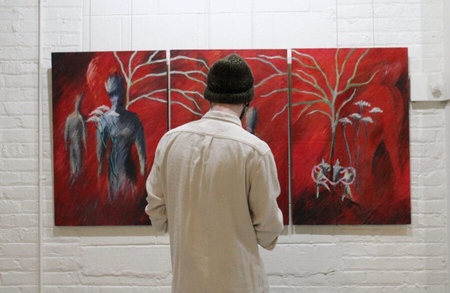 A person wearing a beanie and light shirt stands in front of a three-panel abstract painting with red background and human-like figures.
