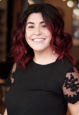 Jacqueline Valdez has black and red curly hair, and is smiling at the camera, wearing a black blouse with black lace sleeves.