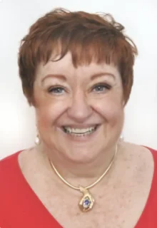 Jean Cozier has short red hair, is wearing a pendant necklace and a red top, and is smiling while standing in front of a white background.