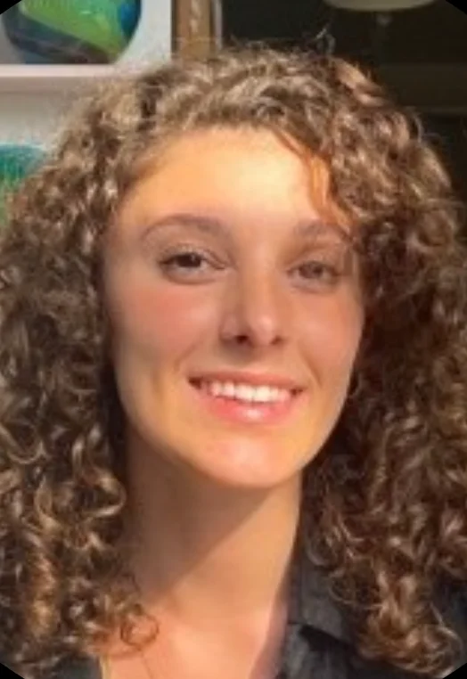 Jules Passell has curly brown hair, is in sunlight in front of ceramic pieces, and is smiling at the camera.