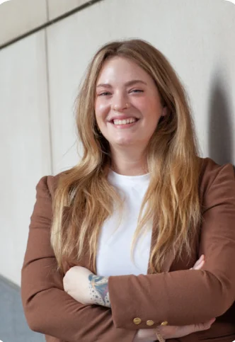 Laura Kinter has long blonde hair and stands with their arms crossed, leaning against a wall, wearing a brown blazer and white shirt.