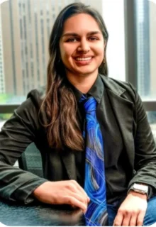 Nisha Kishore is seated in a chair in front of an office window, wearing a bright blue tie and green collared shirt while smiling at the camera.
