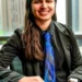 Nisha Kishore is seated in a chair in front of an office window, wearing a bright blue tie and green collared shirt while smiling at the camera.