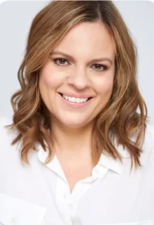 Woman with shoulder-length light brown hair smiles at the camera, wearing a white button-up shirt against a plain light background.