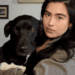 A woman with long dark hair holds a black dog with white markings in front of framed artwork on a wall.