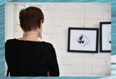 A person with short hair and earrings views two framed black-and-white artworks on a white brick wall in an art gallery.