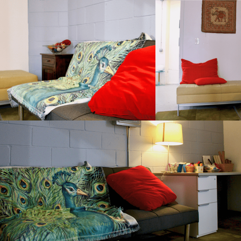 A small living area with a grey futon covered by a peacock-patterned throw and red pillow, a side chair with a red pillow, and a desk with lamp and art supplies.
