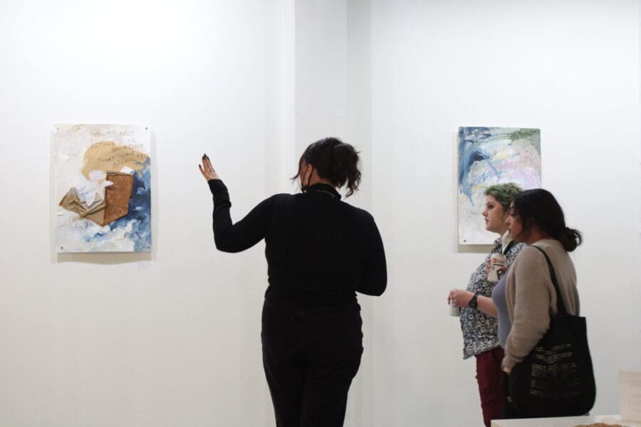 Three people stand in an art gallery, with one gesturing toward two mixed-media paintings displayed on a white wall.