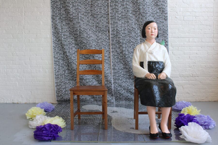 A life-size seated female statue in traditional dress sits next to an empty wooden chair, surrounded by paper flowers, against a patterned backdrop and white brick wall.