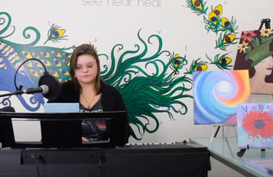 A person sits at a keyboard with a microphone, surrounded by colorful paintings and a peacock mural on the wall behind them.