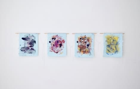 Four abstract mixed media artworks on light blue fabric are hung in a row on a white wall, each featuring varied colors, patterns, and circular shapes.