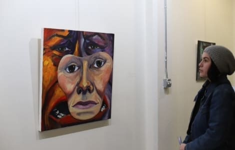 A woman in a gray beanie looks at a colorful abstract painting of a face on display in an art gallery.
