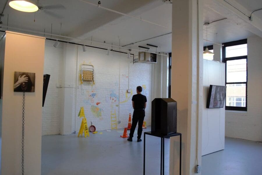A person stands in an art gallery facing a wall with colorful drawings, construction cones, and various objects mounted on it. Artworks are displayed on adjacent walls and stands.