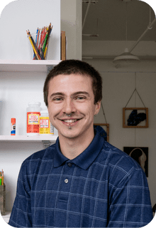 A young man with short brown hair and a goatee, wearing a blue plaid shirt, smiles in an art studio with art supplies and paintings in the background.