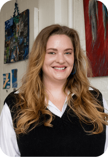 A woman with long, wavy blonde hair smiles while standing indoors in front of colorful artwork on the walls. She wears a white shirt and a black sweater vest.