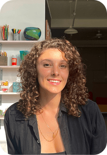A woman with curly hair smiles at the camera in a well-lit room with shelves holding art supplies and decor in the background.
